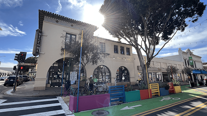 A cream, Spanish-style building with a tree framing its right side and the sun setting behind it. Building reads "SLO Chamber of Commerce Visitor Center". There is a colorfully painted bicycle parking station on the ground in front of the building.